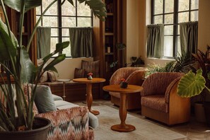 A cozy living room with a sofa, armchair, and a small table with a vase. There are large windows with curtains, and various plants around.