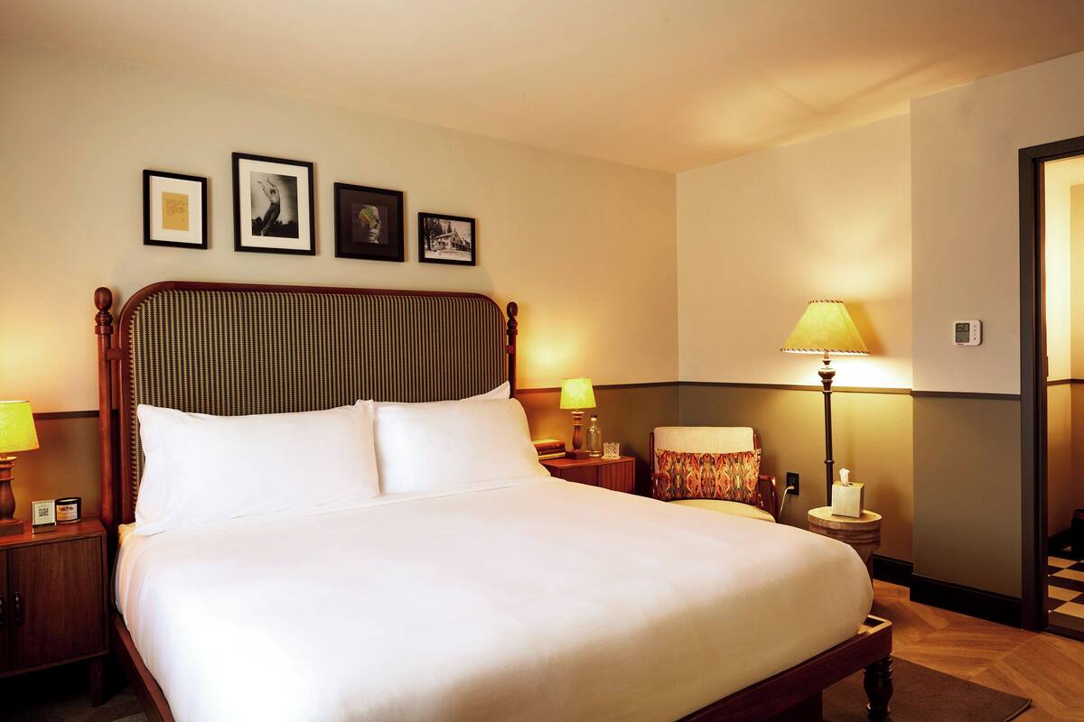 A neatly made bed with white linens, a wooden headboard, and a nightstand with a lamp. There are framed pictures on the wall above the bed.