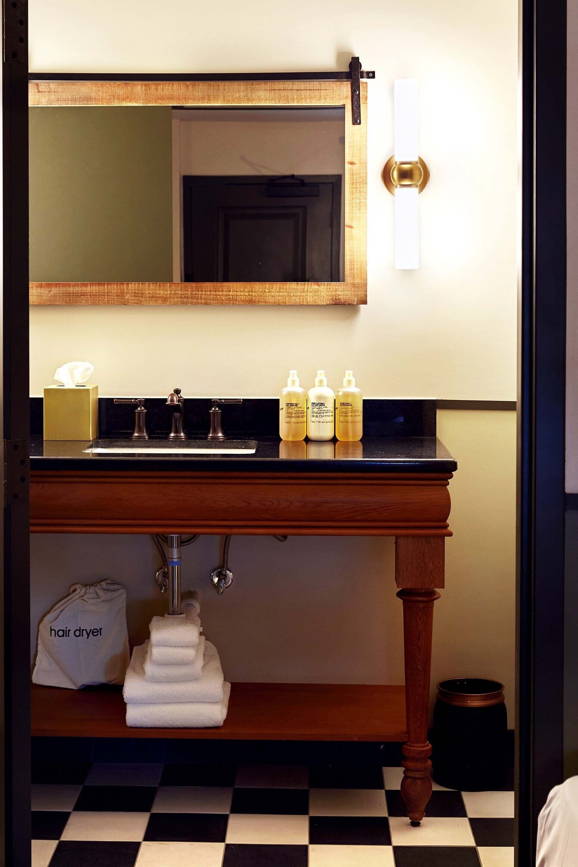 A bathroom with a wooden vanity, a mirror, and a sink. There are toiletries on the counter and a hair dryer on the floor.