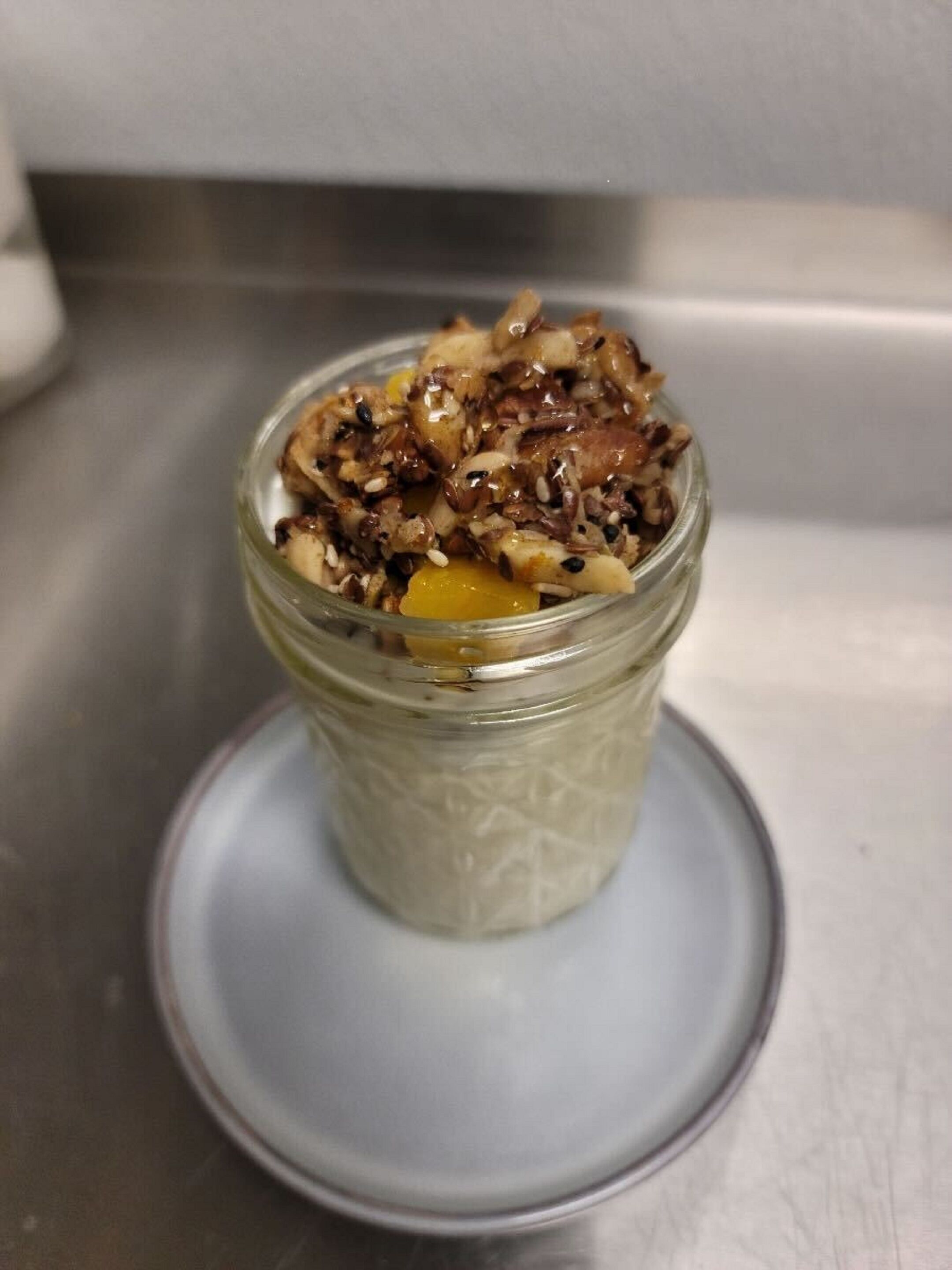 Un bocal en verre contenant un dessert crémeux, garni de noix et de fruits secs, posé sur une assiette blanche sur une surface en acier inoxydable.