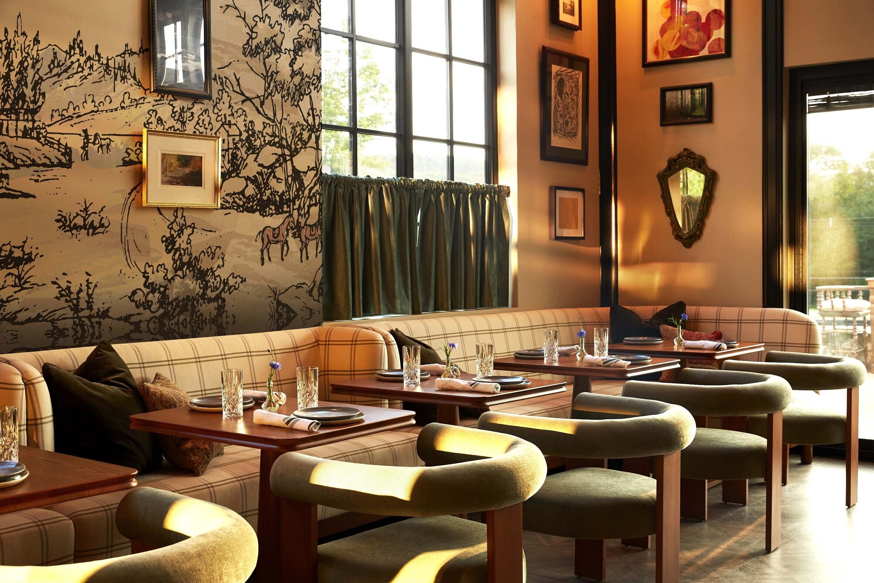 A cozy restaurant with a plaid sofa and wooden tables, framed artwork on the walls, and a large window with curtains.