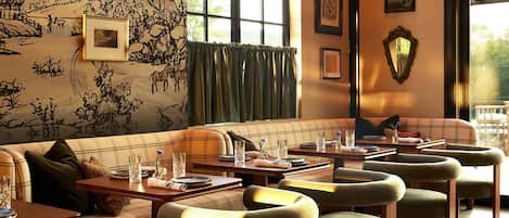 Um restaurante acolhedor com sofá xadrez e mesas de madeira, obras de arte nas paredes e uma grande janela com cortinas.