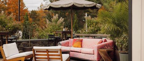 An outdoor seating area with a wooden lounge chair, a cushioned sofa, and a patterned umbrella.