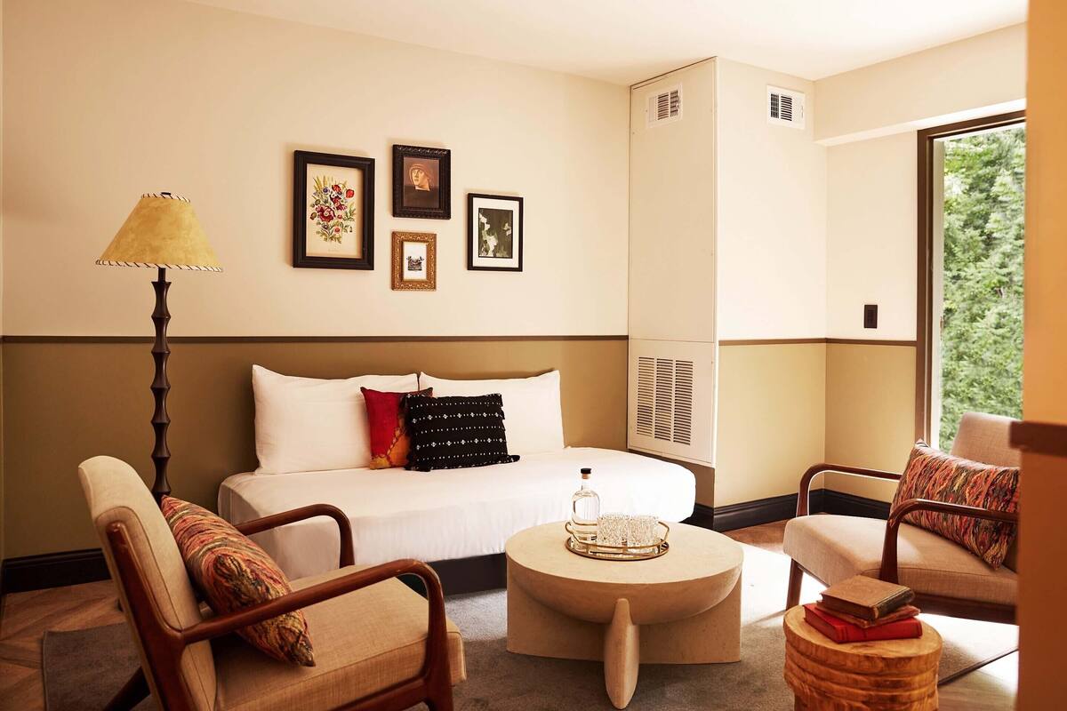 A hotel room with a bed, two armchairs, a round coffee table, and a lamp. There are framed pictures on the wall and a window with a view of greenery.