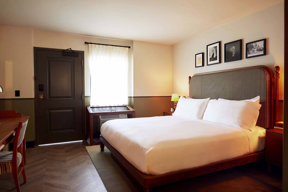 A hotel room with a large bed, a desk, and a chair. There are framed pictures on the wall and a window with curtains.