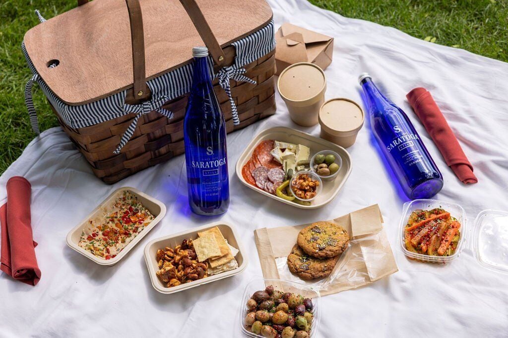 Un montaje para picnic con una cesta de madera, recipientes de comida, bebidas y una manta sobre el césped.