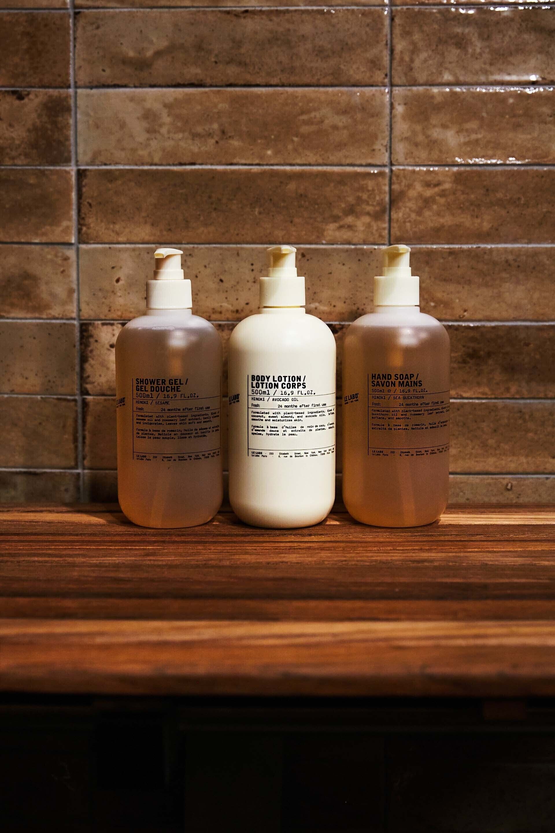 Three bottles of shower gel, body lotion, and hand soap on a wooden surface against a tiled wall.