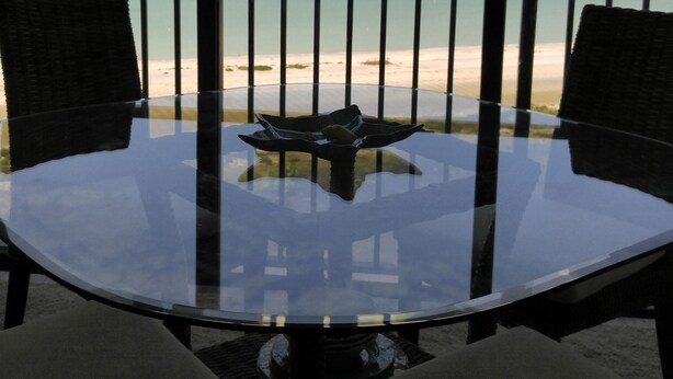 A glass table with two chairs, overlooking a beach with a clear sky and calm sea. - Gorgeous beachfront studio penthouse! (Clearwater Beach)