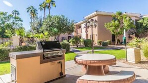 Un área de cocina exterior con una parrilla y una mesa redonda, rodeada de jardines bien cuidados y un edificio al fondo.