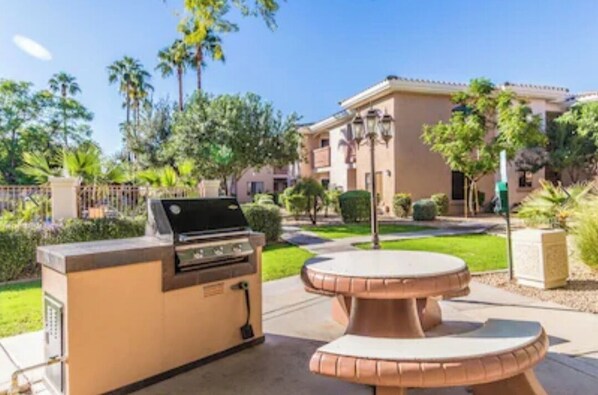 An outdoor kitchen area with a grill and a round table, surrounded by well-maintained landscaping and a building in the background.