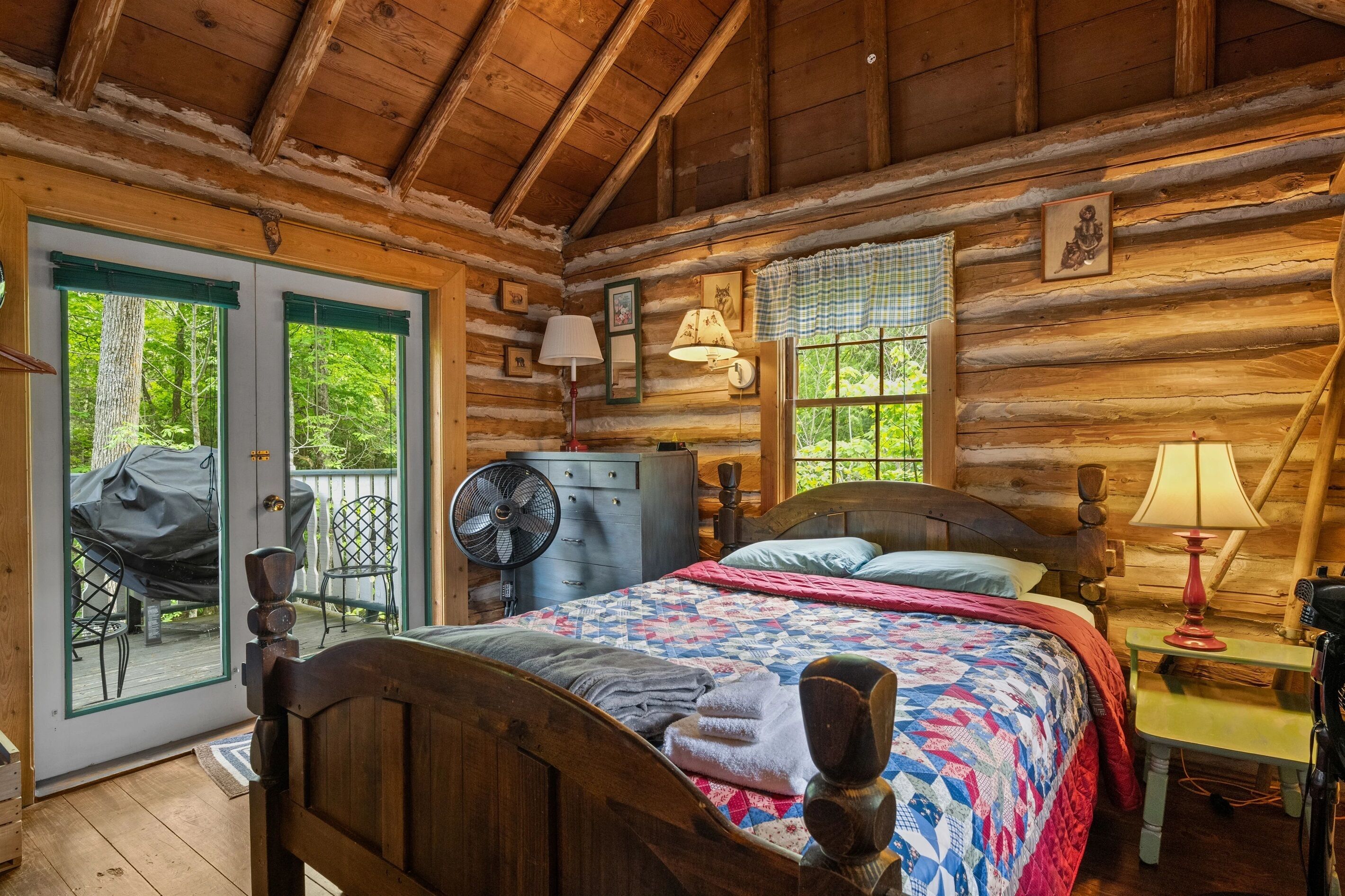 A log cabin bedroom with a bed, two wall-mounted lamps, a fan, and a view of the outdoors.