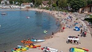 Una playa con kayaks y canoas alineadas en la orilla, un pequeño muelle con barcos, y un edificio con techo inclinado.