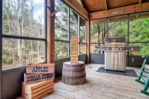 Une terrasse en bois avec un barbecue, des caisses en bois étiquetées « POMMES » et « SUCRES », et un panneau indiquant « LES MONTAGNES APPELLENT ».