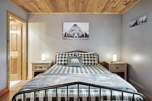 Une chambre avec un plafond en bois, un lit recouvert d’une courtepointe à carreaux, deux tables de chevet avec des lampes et un tableau représentant un ours accroché au mur.
