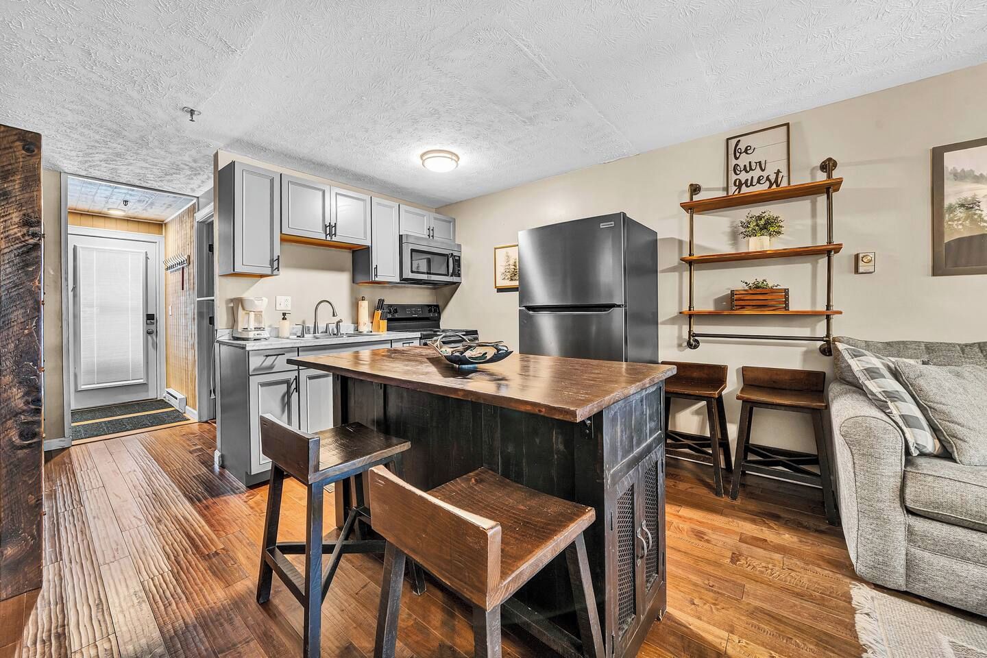A compact kitchen with a bar area, wooden flooring, and a grey sofa.
