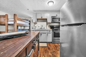 A compact kitchen with a wooden counter, stainless steel appliances, and a bunk bed in the background. - 23 Mountain Crest, Snowshoe, WV - Country Chic (Snowshoe)