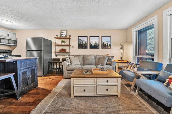 A living room with a sofa, coffee table, and a kitchen area with a refrigerator and microwave.