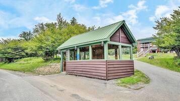 A small wooden structure with a green roof, possibly a restroom or information booth, situated beside a paved path with a grassy area and trees in the background.