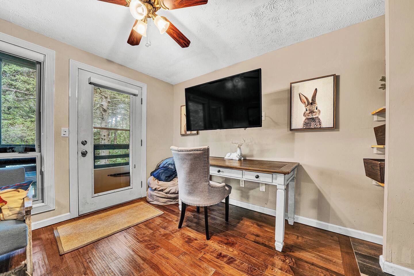 A room with a wooden floor, a desk with drawers, a chair, a television mounted on the wall, and a door leading to an outdoor area.