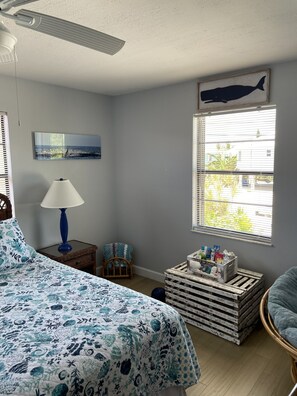 Un dormitorio con cama, mesita de noche, lámpara, silla, ventana con persianas y una obra de arte colgada en la pared.
