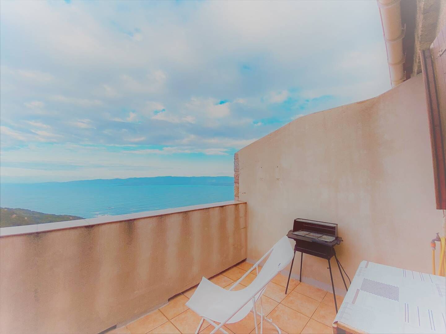 Un balcon avec un barbecue, des chaises blanches et une vue sur la mer.