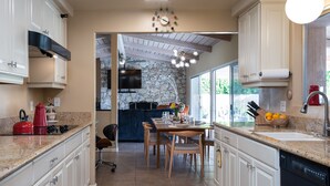 A modern kitchen with white cabinets, a granite countertop, and a stone backsplash. There is a dining area with a wooden table and chairs, and a large window providing natural light. - Fully furnished Midcentury Home, 3 Bd/Den, Views, Best So PD Location (Palm Desert)