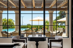 A room with a large window overlooking a pool area, furnished with wicker chairs and a table with chess pieces. - Oceanfront Island View Gated Santa Barbara Villa (Santa Barbara)