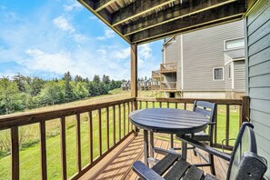 A wooden deck with a table and chairs, overlooking a grassy area and a building with multiple balconies. - 58 Powderidge, Snowshoe, WV - Cozy Chalet (Snowshoe)