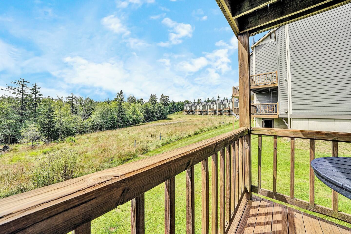 A wooden deck with a view of a grassy field and trees, leading to a building with a balcony.