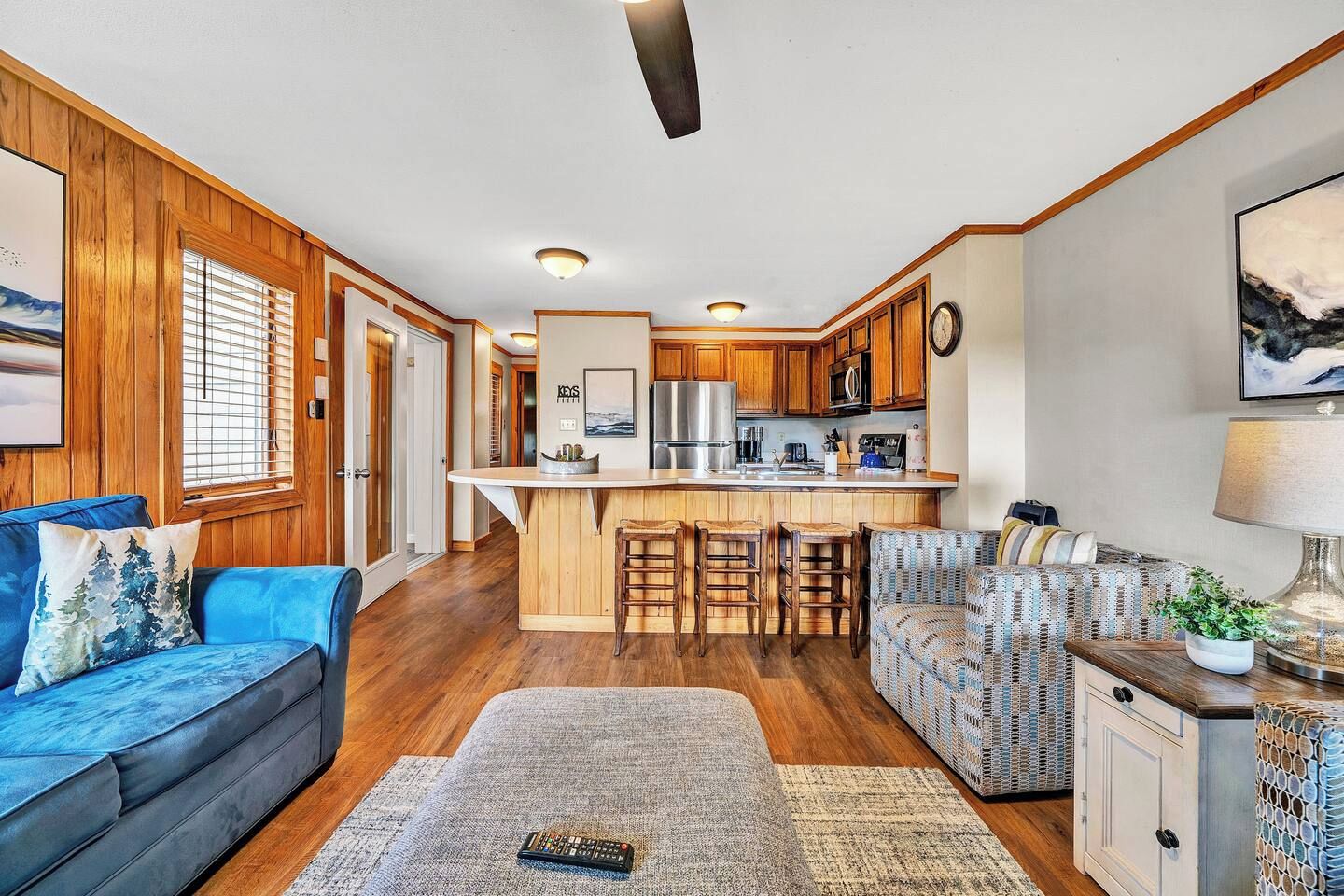 A modern kitchen with wooden cabinets and a central island, a living area with a blue sofa and patterned armchair, and a dining area with a wooden table and chairs.
