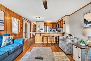 A modern kitchen with wooden cabinets and a central island, a living area with a blue sofa and patterned armchair, and a dining area with a wooden table and chairs.