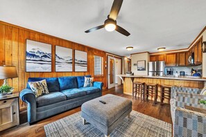 A modern living room with a blue sofa, a grey ottoman, and a wooden ceiling fan.
