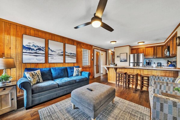 A modern living room with a blue sofa, a grey ottoman, and a wooden ceiling fan. - 58 Powderidge, Snowshoe, WV - Cozy Chalet (Snowshoe)