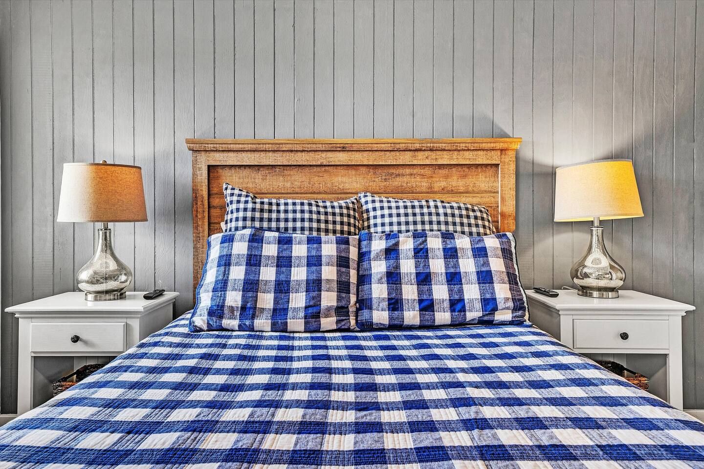 Ein Bett mit einer blau-weißen, karierten Tagesdecke, zwei passende Kissen, ein hölzernes Kopfende und zwei Nachttische mit Lampen.