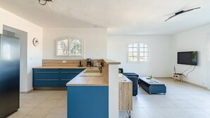 Una cocina moderna con armarios azules, encimera de madera y un lavaplatos integrado. Hay una zona de comedor con un sofá azul y una mesa de centro. Un televisor está montado en la pared, y hay ventanas que proporcionan luz natural.