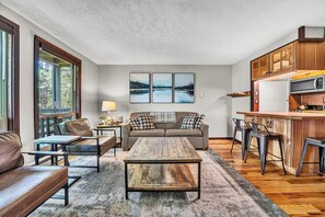 A living room with a sofa, coffee table, and chairs, a kitchen with a microwave and bar stools, and a large window with a view of trees. - 24 Mountain Crest, Snowshoe, WV - Rustic Retreat (Snowshoe)
