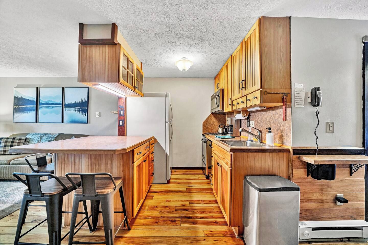 A compact kitchen with wooden cabinets, a refrigerator, a microwave, and a coffee maker. There is a bar area with stools and a small table.