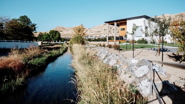A modern building with a flat roof and large windows, situated next to a river with a stone pathway leading to it. - Modern Apt w/ Private Entry & Patio - Mtn Views (Smithfield)