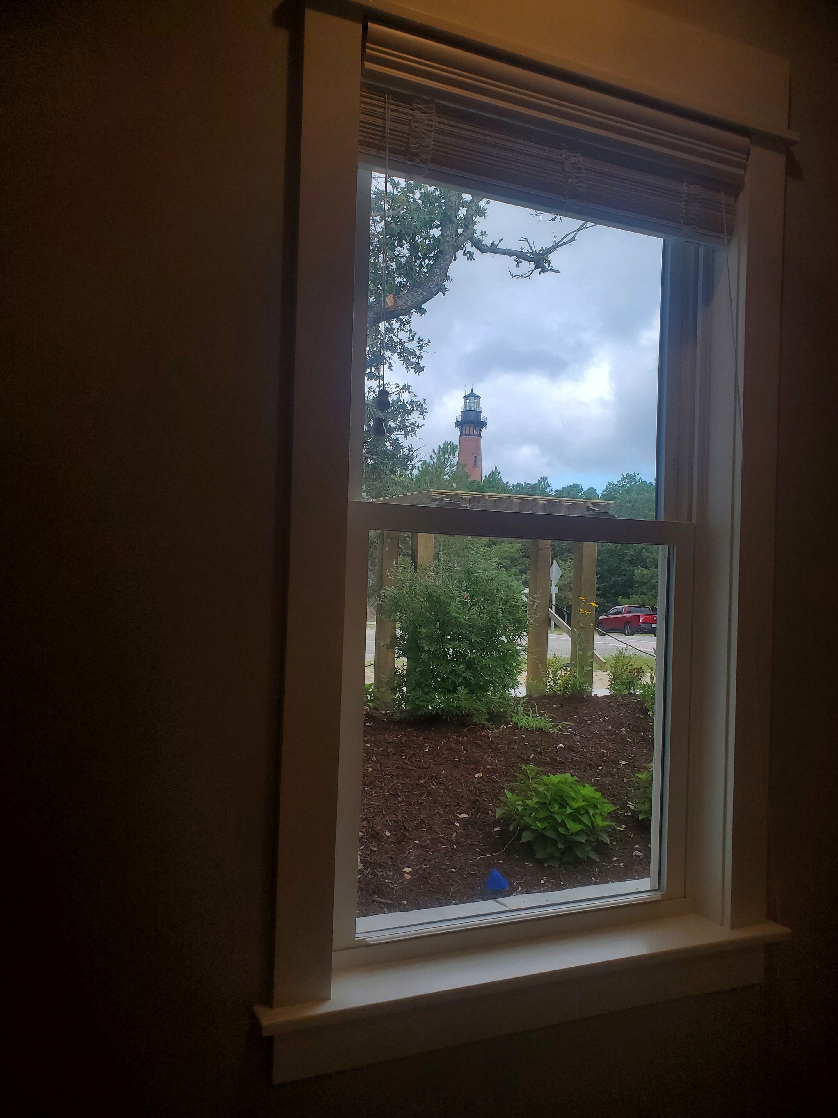 A window with a view of a lighthouse, trees, and a parking area.