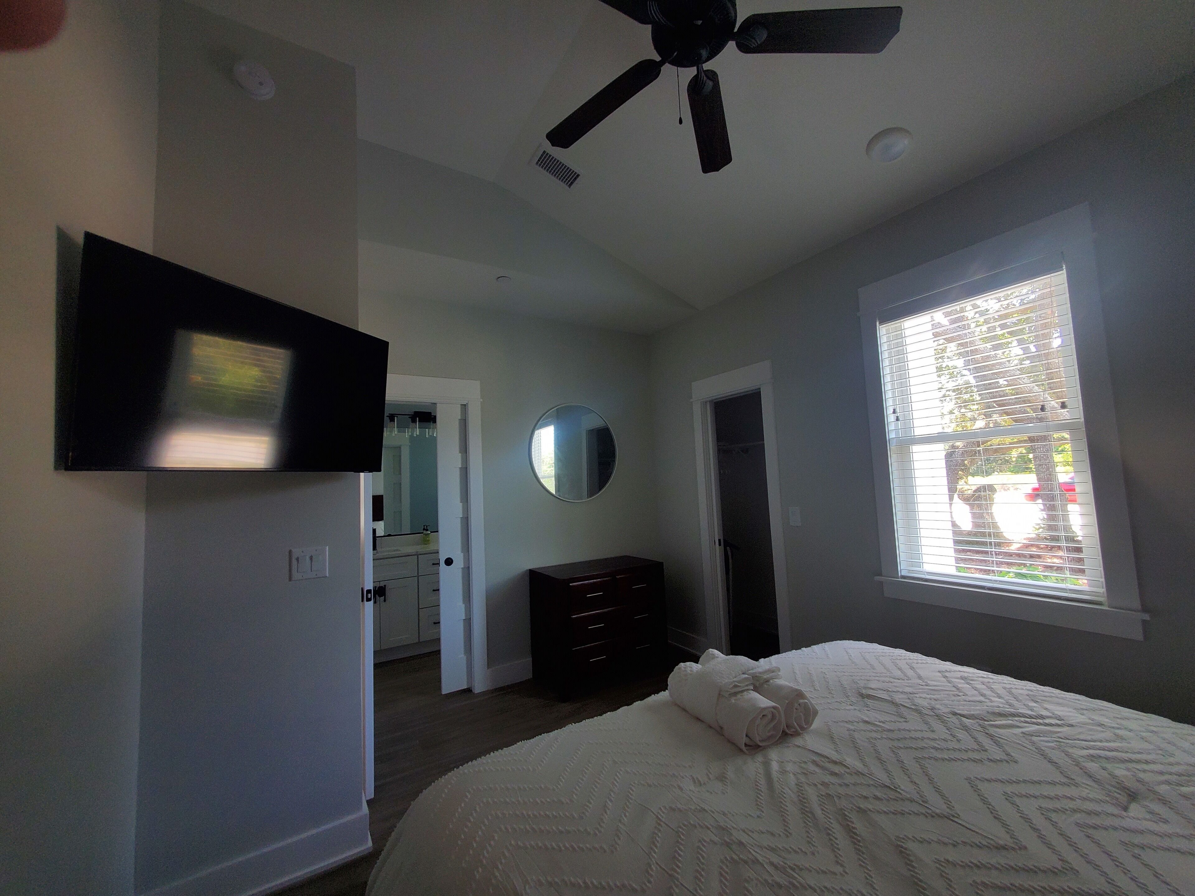 A bedroom with a bed, a ceiling fan, a TV, a mirror, and a window.