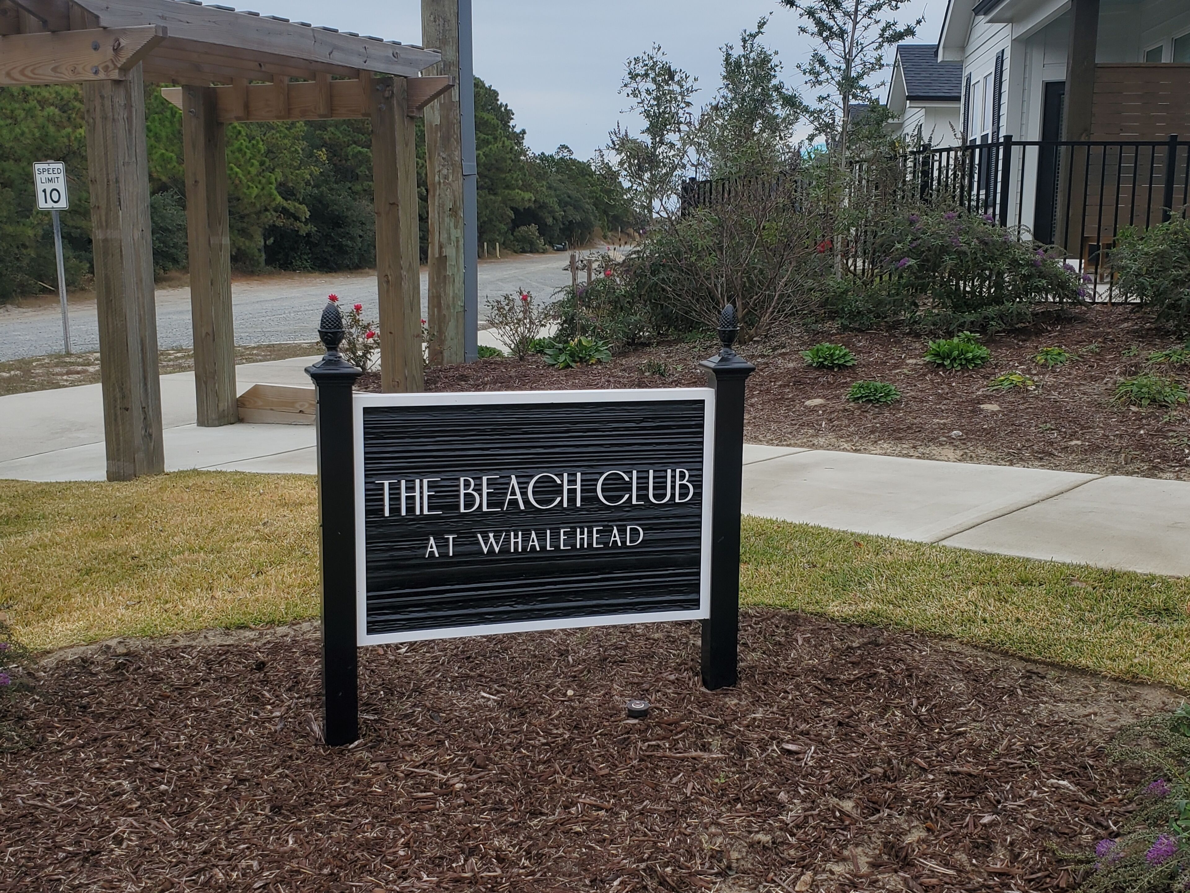 A sign for 'The Beach Club at Whalehead' with a wooden pergola and a speed limit sign in the background.