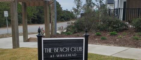 A sign for 'The Beach Club at Whalehead' with a wooden pergola and a speed limit sign in the background.