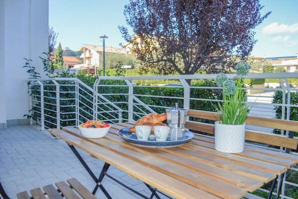 An outdoor dining area with a wooden table, a bowl of fruit, a croissant, and a coffee machine.