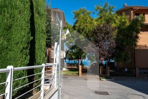 Une entrée fermée par un portail et agrémentée d’une rampe métallique, un chemin menant à un bâtiment doté d’un balcon, et une vue sur la mer.
