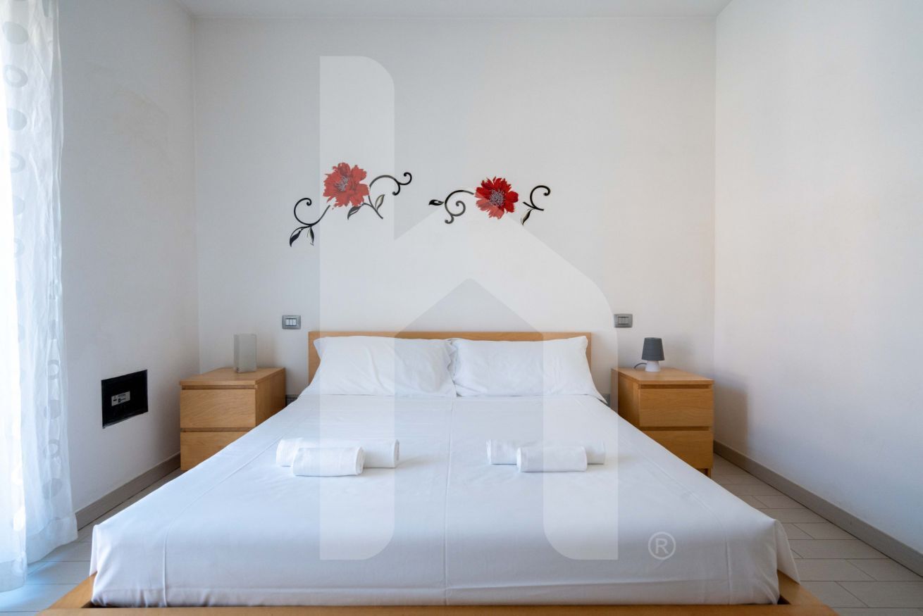 A neatly made bed with white linens, two bedside tables with lamps, and a wall decoration of red flowers.