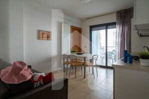 A modern living room with a sofa, a dining table with chairs, a kitchen area, and a sliding glass door leading to an outdoor balcony.