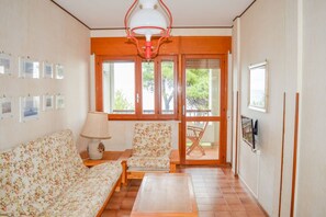 Ein Wohnzimmer mit einer Couch, einem Sessel und einem Tisch mit einer Lampe. Fenster bieten Blick auf Grünflächen.