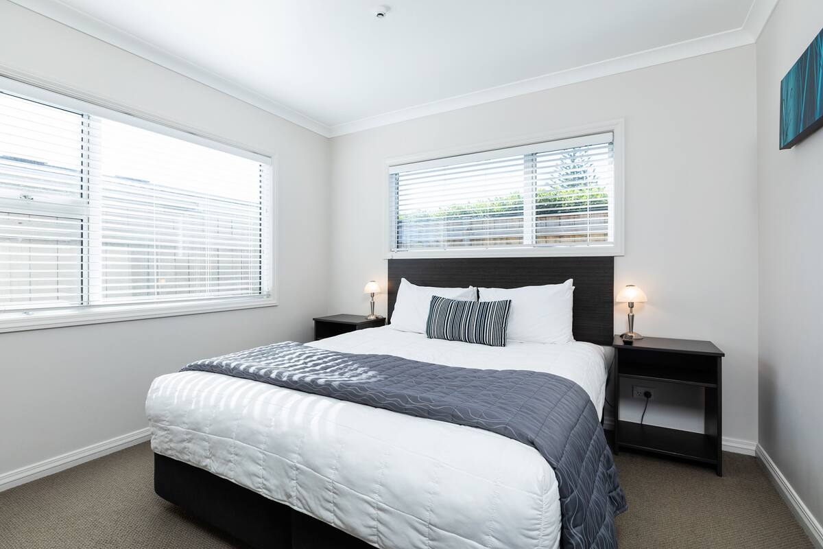 A bedroom with a bed, two bedside tables, a headboard, and two windows with blinds.