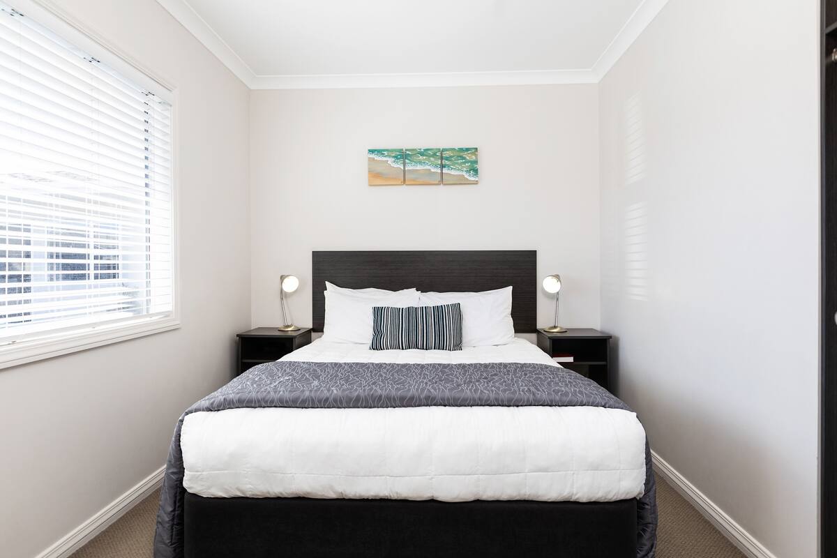 A neatly made bed with a white and gray comforter, a dark headboard, and a single pillow. There is a small bedside table with a lamp on each side of the bed. A painting hangs on the wall above the bed.