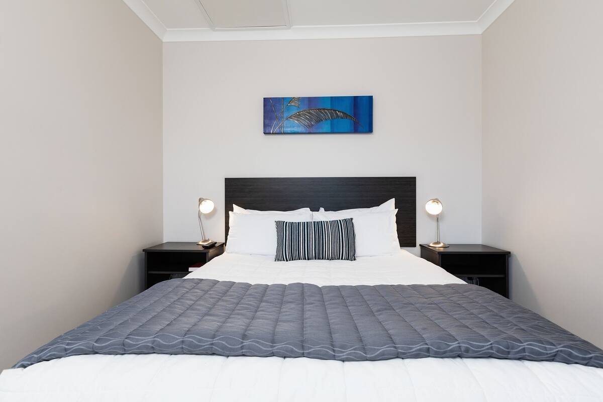 A neatly made bed with a gray quilt, a white pillow, and a striped pillow. There is a blue painting on the wall above the bed. Two bedside tables with lamps are on either side of the bed.